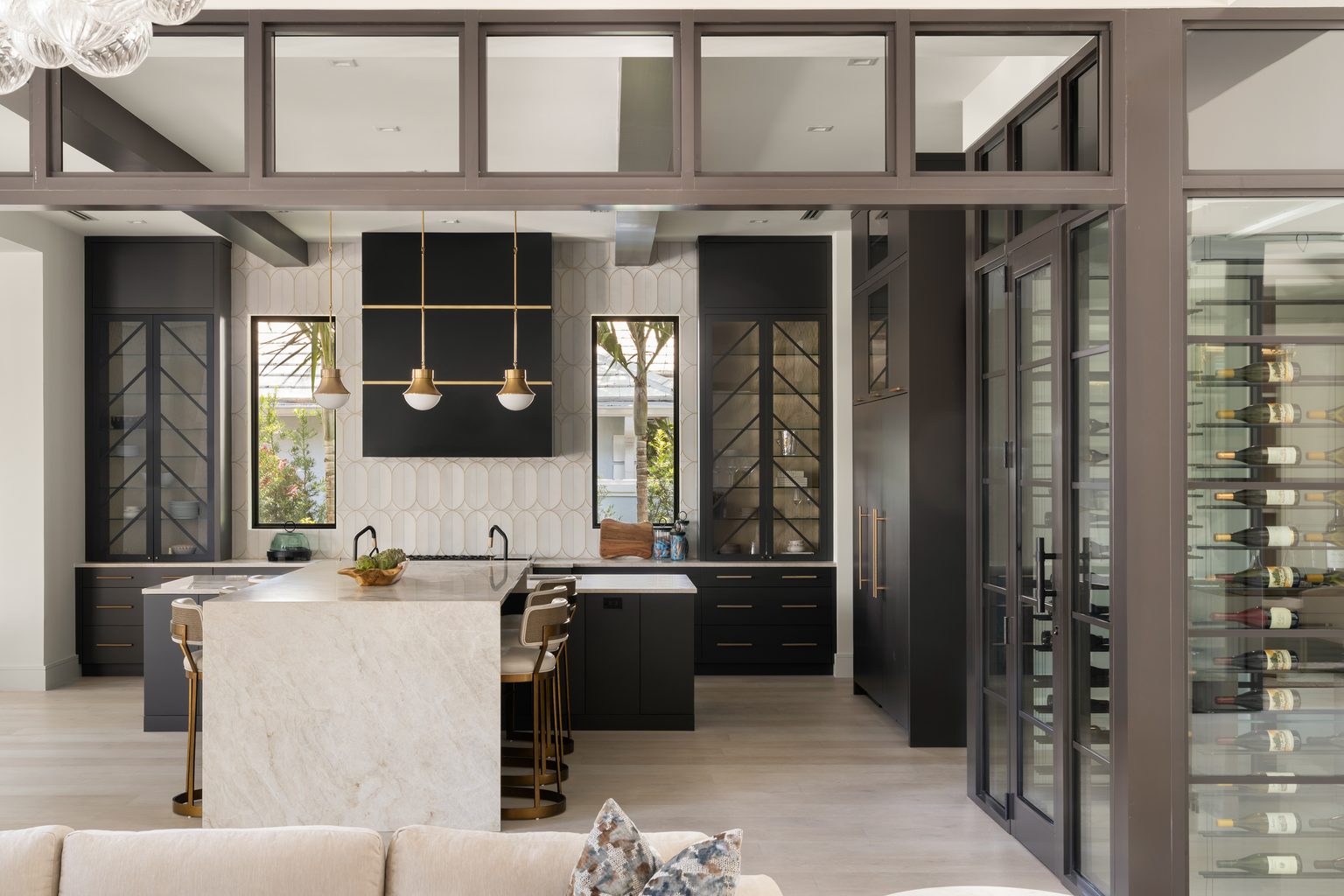 A modern style kitchen and wine cellar with glass doors. In the kitchen, there's a perpendicular Taj Mahal island, matte black cabinetry, panel-ready appliances, brass accents, and a bronze frame around the ceiling and wine cellar.